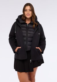 Giacca a vento nera con cappuccio e tasche laterali, dotata di chiusura frontale con zip e texture trapuntata, indossata sopra un cappotto aderente nero.