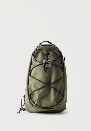 Olivengrøn Carhartt rygsæk med sorte justerbare bungee snore på fronten og top bærehåndtag mod en ensfarvet lys baggrund.