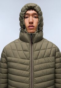 Giacca imbottita verde oliva con motivo a rombi, cappuccio alto, e chiusura frontale con zip, dotata di cordoncini regolabili e finitura testurizzata.