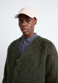 Πράσινο κερκίδα με υφή, με V-λαιμό και κουμπιά, τοποθετημένο πάνω από ένα μπλε και καφέ καρό πουκάμισο, με μια μπεζ καπέλο μπέιζμπολ από πάνω.