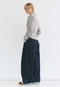Camicia a righe con bottoni abbinata a pantaloni in denim blu scuro, larghi, con risvolti e tasche posteriori. Il modello è in posa di lato su uno sfondo bianco.