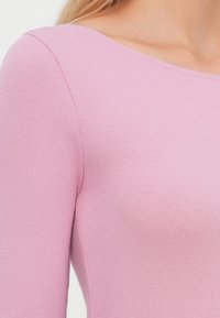 Nærbilde av en person som har på seg en ribbet, lys rosa, langermet topp med rund hals.