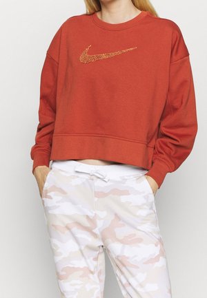 Vrouw die een roestoranje sweatshirt draagt met een glinsterend Nike-logo en wit-roze beige camouflagebroek met trekkoord, handen in de zakken, staand tegen een witte achtergrond.