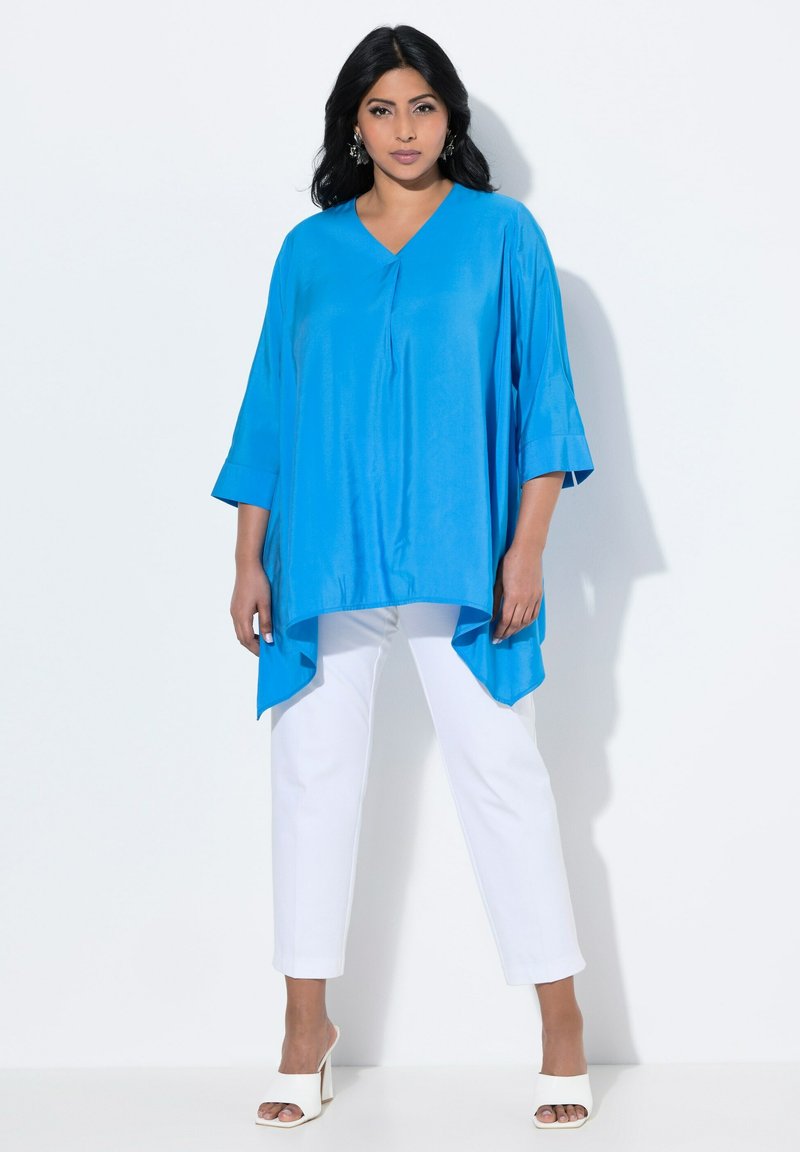 Top in satin blu con scollatura a V e maniche a tre quarti, abbinato a pantaloni bianchi su misura e sandali bianchi con tacco a blocco.