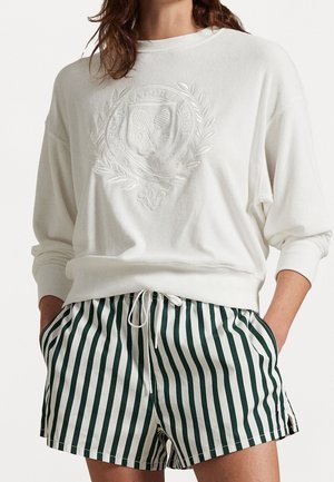 White sweatshirt with tennis emblem paired with green and white vertical striped drawstring shorts, hands tucked in pockets.
