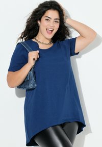 Maglietta blu a maniche corte con orlo curvo e vestibilità rilassata, abbinata a leggings neri e una piccola borsa in denim. Gioielli con catena d'argento come dettagli.