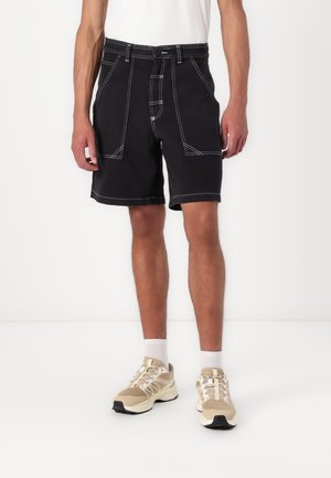 Black shorts with white stitching and a high waist. Features two front pockets and a belt loop. Paired with beige athletic shoes.