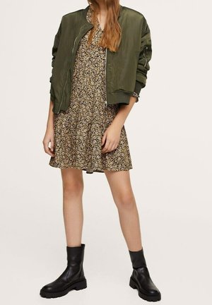 Vrouw draagt een groene bomberjas over een korte bloemenjurk, gecombineerd met zwarte grove enkellaarsjes, staand met de benen iets uit elkaar.