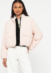 Veste courte rose clair avec fermeture éclair à l'avant, deux grandes poches à l'avant et poignets élastiqués. Haut texturé noir en dessous. Pantalon blanc.