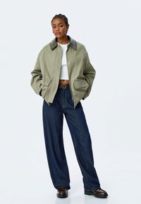 Olivgrön jacka med svart krage, knäppt framtill och två sidofickor; kombinerad med vida mörkblå jeans och svarta skor.