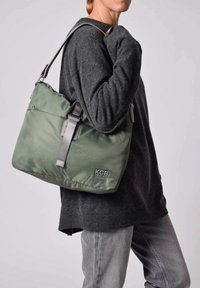 Bolso vegano de color verde oliva hecho de material suave, con una correa gris y un logotipo en la parte frontal. El bolso tiene una forma relajada y flexible con bordes redondeados.