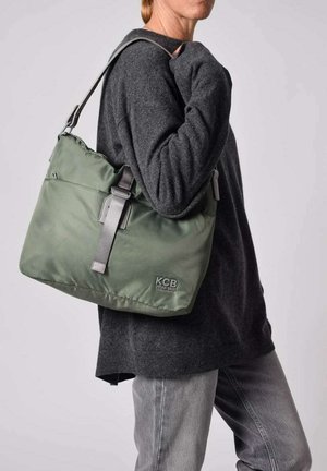 Bolso vegano de color verde oliva hecho de material suave, con una correa gris y un logotipo en la parte frontal. El bolso tiene una forma relajada y flexible con bordes redondeados.