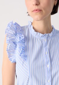 Hellblau gestreifte Bluse mit Rüschenschößen und rundem Halsausschnitt, mit einer Knopfleiste vorne und weicher Stofftextur.