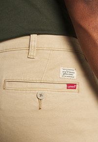 Pantaloni in cotone beige con tasca posteriore con chiusura a bottone e un'etichetta in tessuto bianca con "Levi Strauss" e un'etichetta rossa "Levi's".