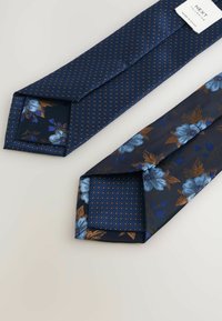 Divi divi zili kaklasaiti, viens ar maziem oranžiem punktiem, otrs ar gaiši ziliem un brūniem ziedu rakstiem, redzams uzraksts "Next Tailoring".