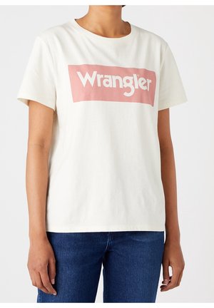 Osoba nosząca białą koszulkę z krótkim rękawem z różowym prostokątnym logo z napisem „Wrangler” i niebieskie dżinsy na jednolitym tle.