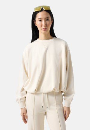 Kvinde med langt sort hår iført en løs cremefarvet sweatshirt, matchende bukser og solbriller med gult skær på hovedet, stående mod en hvid baggrund.