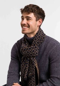 Uomo sorridente con capelli ricci corti che indossa un maglione a coste scuro e una sciarpa geometrica nera e marrone contro uno sfondo semplice.