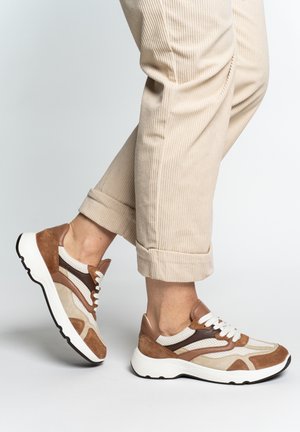 Person wearing beige corduroy pants and brown, white, and tan lace-up sneakers walking on a light gray surface.