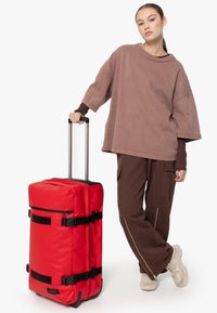 Valise à roulettes rouge avec des sangles et des fermetures éclair noires, dotée d'une poignée robuste et d'une texture en tissu, accompagnée d'une personne vêtue d'une tenue décontractée marron.