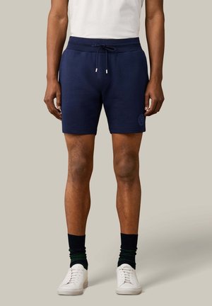 Man draagt marineblauwe short met trekkoord, zwarte sokken, witte sneakers en een wit T-shirt tegen een effen lichtgrijze achtergrond.