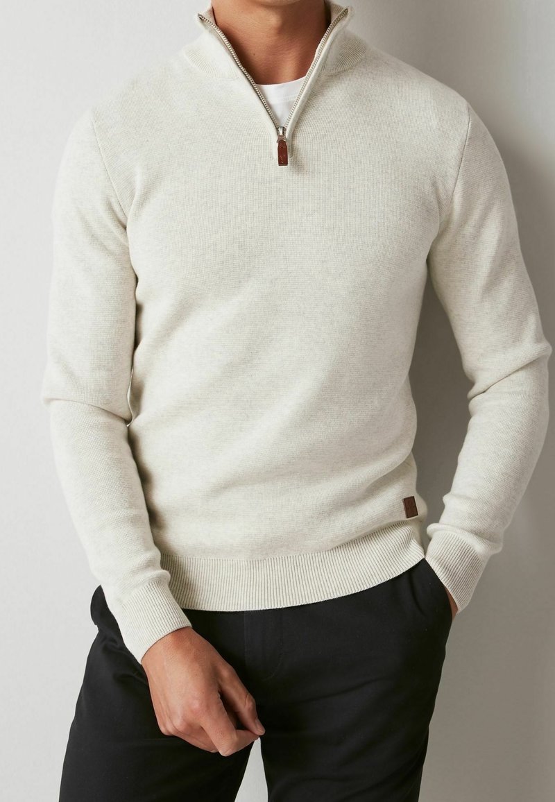 Homme portant un pull en tricot crème à demi-fermeture éclair sur une chemise blanche, avec un pantalon noir, debout avec une main dans la poche devant un fond uni.