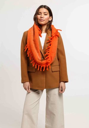 Femme portant un blazer marron, un pantalon blanc et une écharpe à franges orange vif, se tenant devant un fond clair.