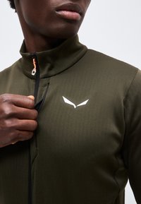 Κοντινό ενός ατόμου που κλείνει το φερμουάρ ενός ελαιόχρωμου μπουφάν με ψηλό γιακά και λευκό γωνιακό λογότυπο στο στήθος.