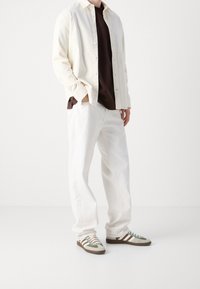Camicia leggera di colore crema sopra una t-shirt marrone, abbinata a pantaloni bianchi a gamba larga e sneakers verde con strisce marroni.