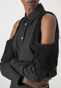Camicia nera con colletto classico, caratterizzata da frange nere lunghe sulle spalle. Tessuto liscio, chiusura a bottoni, tatuaggi visibili sulle braccia.