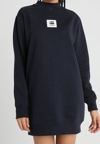 Mörkblå sweatshirtklänning med långa ärmar, ribbade ärmmuddar och en rektangulär vit logotyp på bröstet. Mjuk bomullstextil.