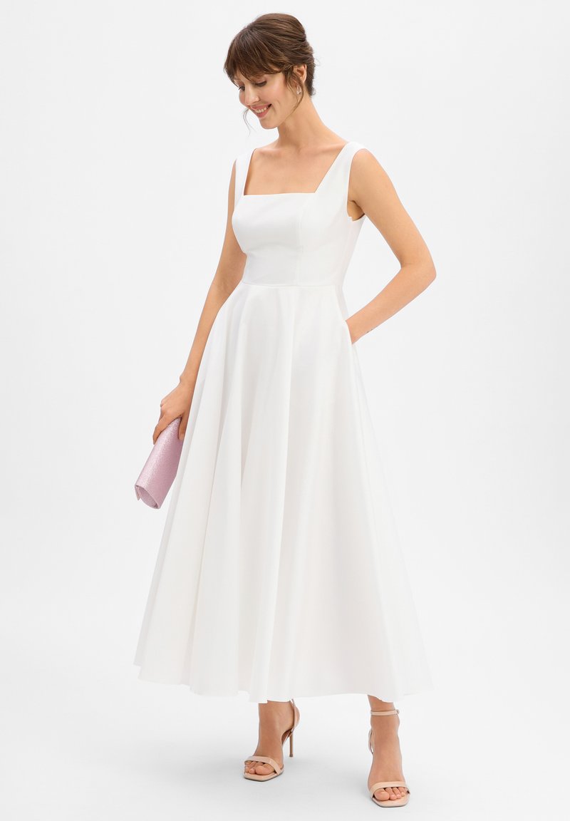 Robe blanche ligne A avec un décolleté carré, de larges bretelles et une jupe fluide. Associée à une pochette rose et des talons nus.