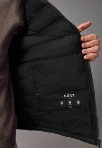Sort polstret jakke med indvendig for, der viser quiltet tekstur, label med "NEXT" og plejesymboler for varme og vandtæthed.