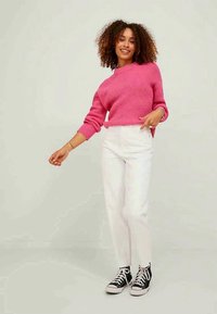 Vaaleanpunainen ribbipaita korkean kauluksen kanssa, yhdistettynä valkoisiin suoriin housuihin ja mustiin korkeavartisiin tennareihin. Seisoen neutraalilla taustalla.