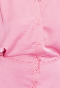 Ljusrosa skjorta med knappar framtill som har en slät yta, sex knappar nedanför och en klassisk krage.