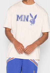 Man som bär en beige T-shirt med blått "MN" och en blå Playboy-kaninlogotyp, ihop med blå shorts med röda och vita ränder samt en gul kaninlogotyp.