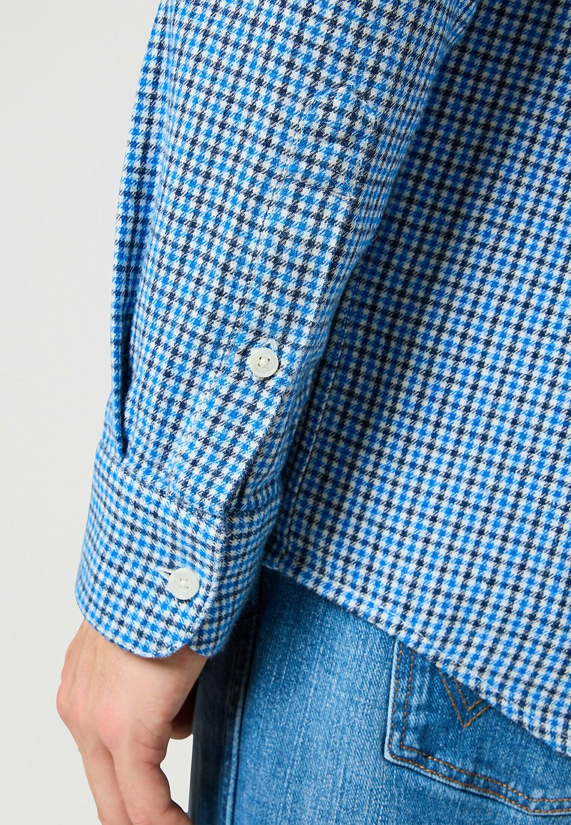 Camicia a scacchi blu e bianca con polsini abbottonati, realizzata in un tessuto texturizzato. Abbinata a jeans blu, mostra un contrasto di colore.