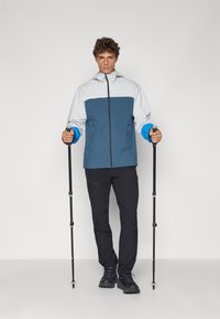Veste de randonnée bleue et blanche avec fermeture éclair à l'avant, poches latérales et manches ajustables. Un pantalon noir et des bâtons de marche complètent la tenue de plein air.