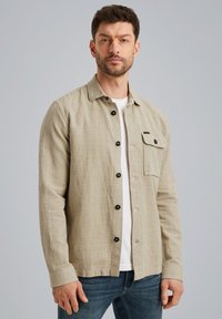 Lichtbeige overhemd met knopen in een textuurstof, voorzien van een borstzak en zwarte knopen, gecombineerd met een effen wit T-shirt en jeans.