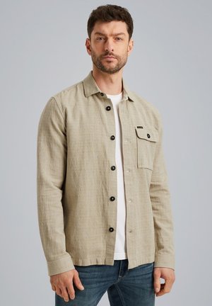 Light beige button-up shirt in textured fabric, featuring a chest pocket and black buttons, paired with a plain white T-shirt and jeans.