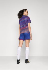 Femme avec une queue de cheval bouclée portant un maillot de sport rayé violet, un short bleu, des chaussettes blanches montantes et des crampons noirs, se tenant contre un mur gris.