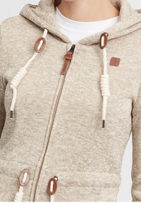 Sudadera beige con cremallera, con cordones blancos de cuerda y detalles de cuero marrón, puesta sobre una camiseta blanca de cuello redondo.
