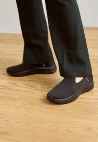 Black slip-on shoes with a textured mesh upper, padded collar, and a solid rubber sole, worn with dark green pants on a wooden floor.