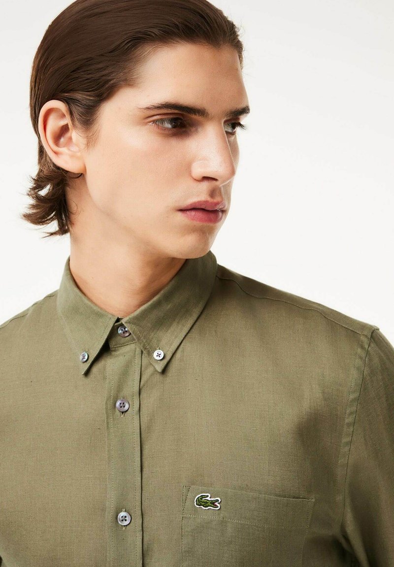 Camicia verde oliva con bottoni, finitura testurizzata, taschino sul petto e logo ricamato. Presenta bottoni grigi e un design colletto classico.