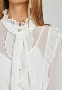 Weiße Bluse mit rüschigem Ausschnitt, zarten Knopfverschluss und fließendem Stoff mit transparenten Details an den Kanten.