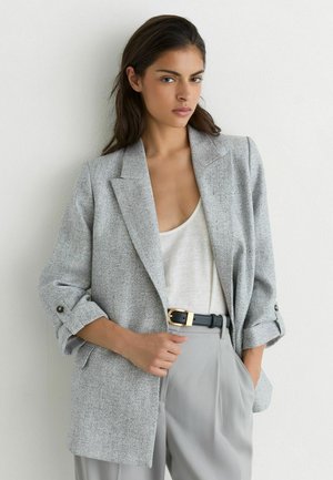 Femme portant un blazer gris texturé sur un haut blanc avec un pantalon gris et une ceinture noire, debout contre un mur blanc.