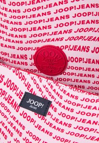 Rdeče in roza vzorčasta tkanina z ponavljajočim se besedilom "JOOP! JEANS", ki vključuje okroglo rdečo oznako in črno etiketo. Teksturirana površina.