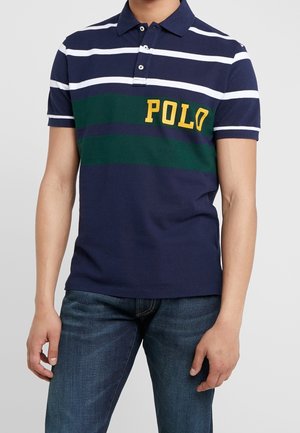 Homme portant un polo rayé bleu marine, vert et blanc avec "POLO" en lettres jaunes, associé à un jean bleu.