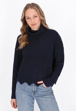 Maglione lavorato a maglia blu navy con altocollo, motivo a trecce e orlo ondulato, abbinato a jeans in denim chiaro.