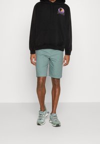 Svart hoodie med grafisk design, kombinerat med ljusgröna shorts och grå träningsskor, som visar en avslappnad passform och avslappnad stil.